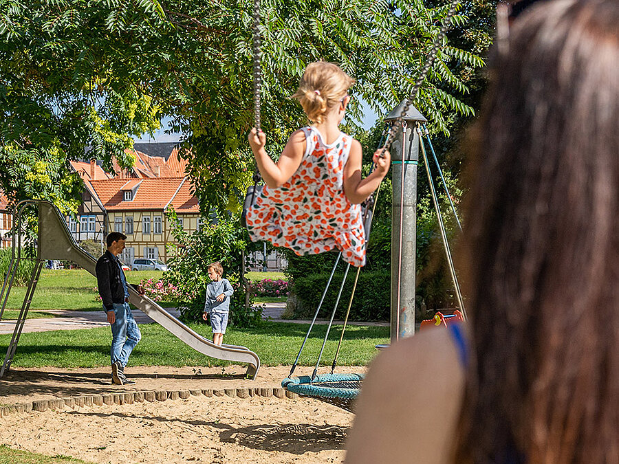Eine Familie auf einem Spielplatz: Im Vordergrund eine Frau mit einem Kind auf einer Schaukel, im Hintergrund ein Mann mit einem Kind an einer Rutsche.