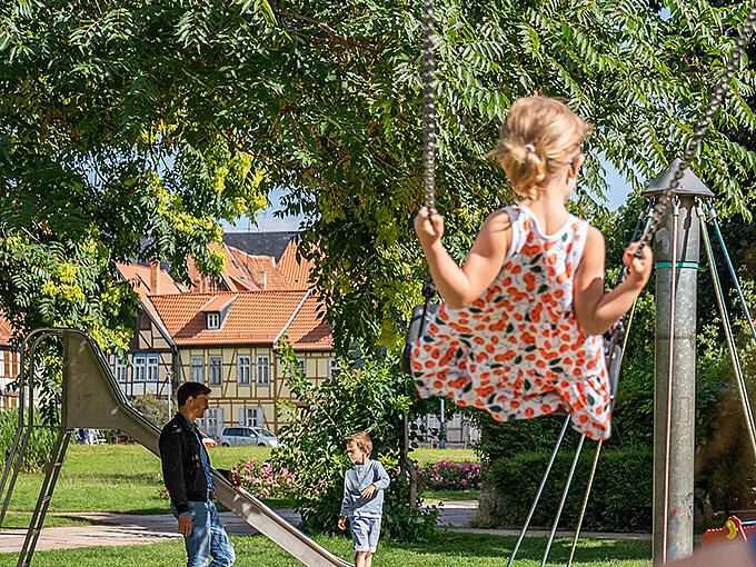 Eine Familie auf einem Spielplatz: Im Vordergrund eine Frau mit einem Kind auf einer Schaukel, im Hintergrund ein Mann mit einem Kind an einer Rutsche.