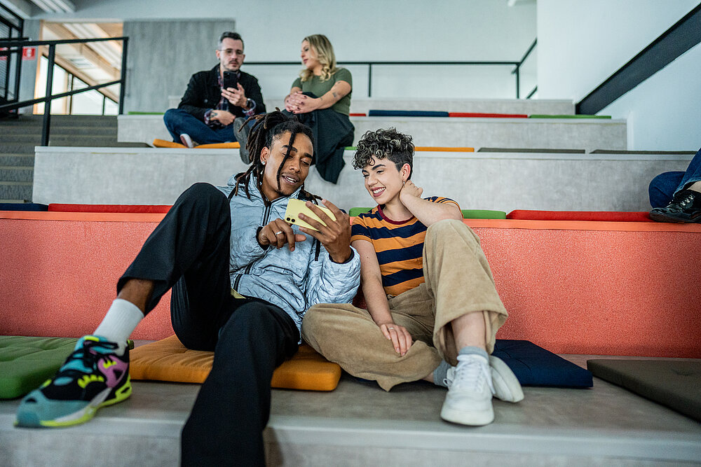 Vier Schüler sitzen auf einer Zuschauertribühne und schauen auf Handys und diskutieren.