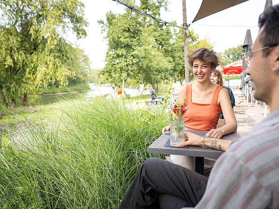 Zwei Personen sitzen mit Getränken an einem Tisch im Park, im Hintergrund ein Fluss.