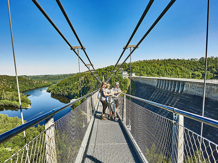 Drei Personen stehen auf einer Hängebrücke über einem Fluss.
